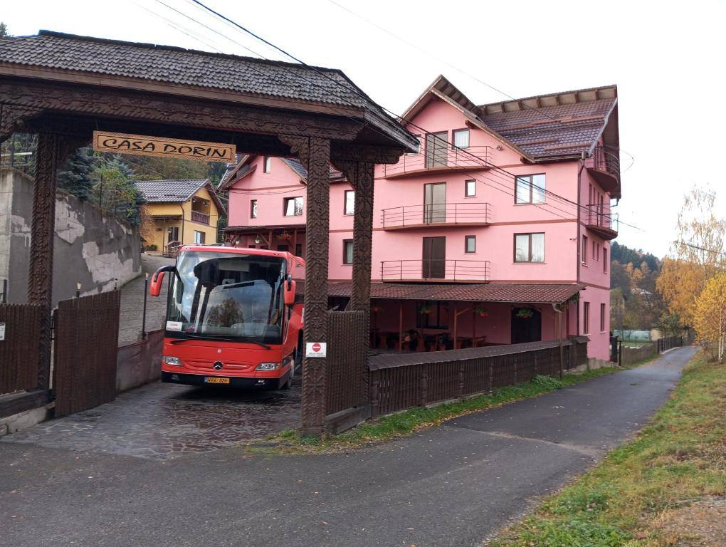 Autocar parcat sub poarta maramureșeană a pensiunii, lângă clădirea roz cu balcoane pe trei niveluri.