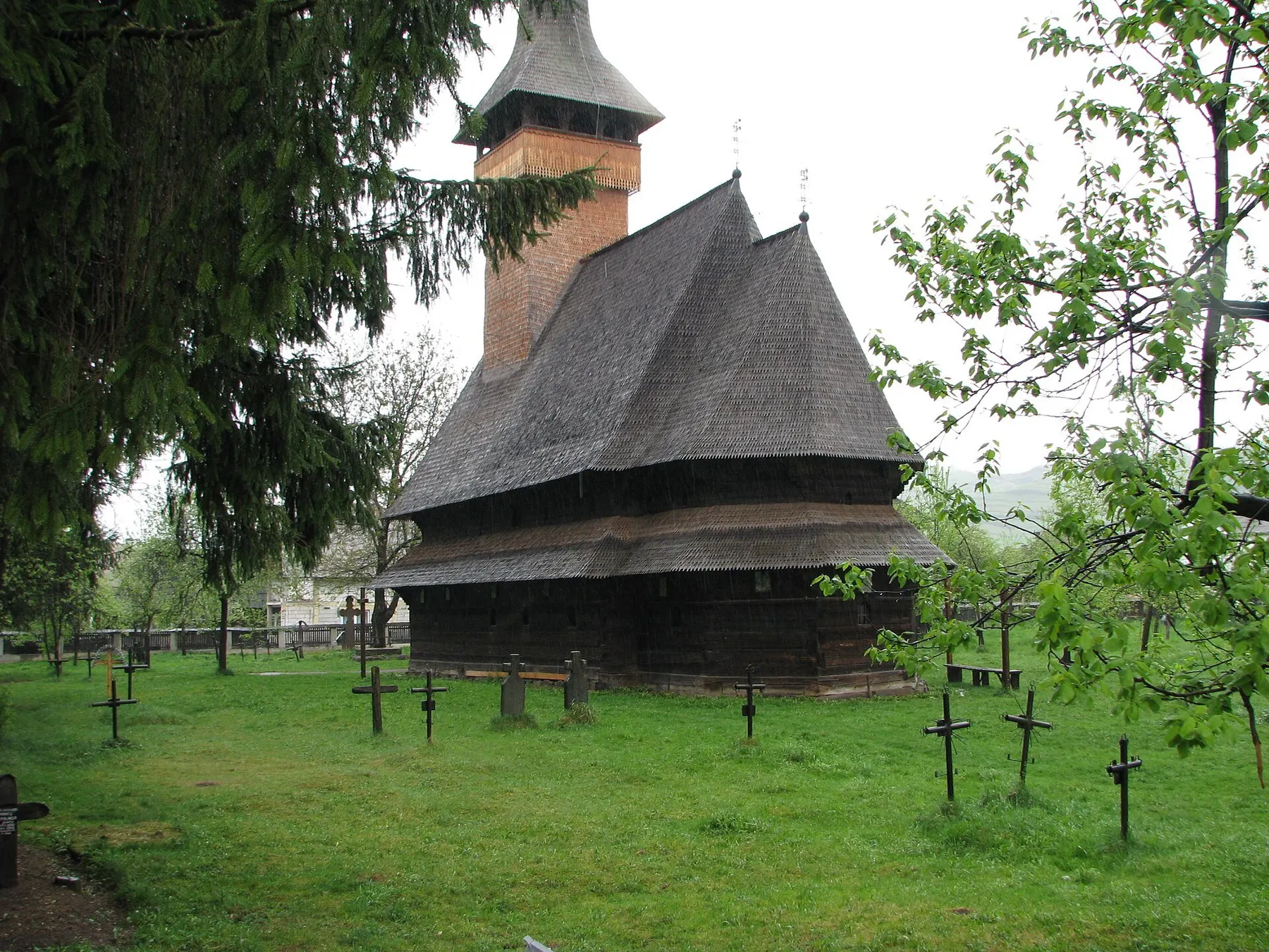 Biserica de lemn din Bogdan Vodă, cu acoperiș în trepte, într-un cimitir cu iarbă și cruci de lemn.