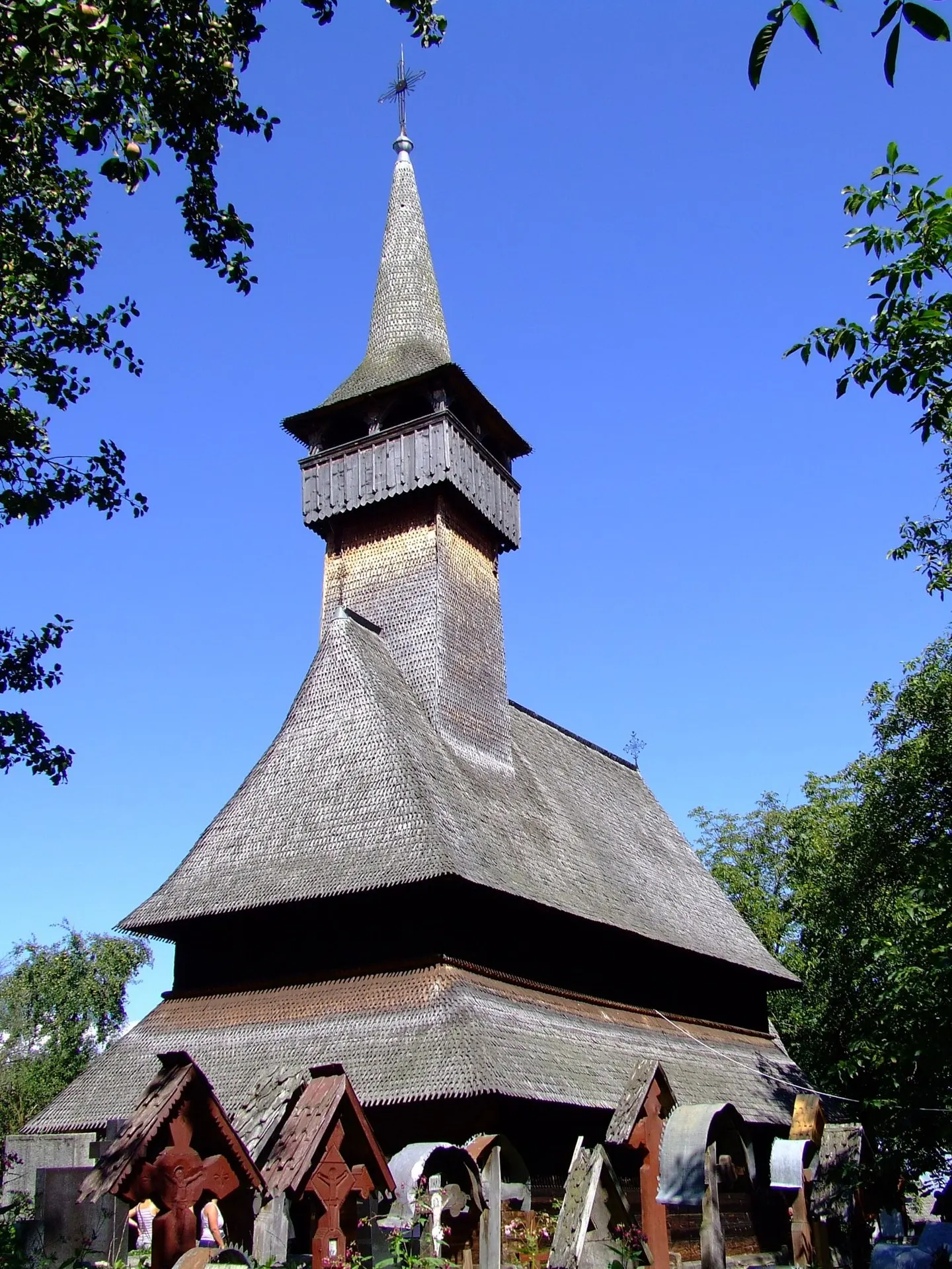 Biserica de lemn din Ieud Deal, cu turn înalt și acoperiș de șindrilă, între cruci vechi.