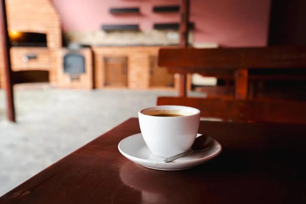 O ceașcă de cafea pe o masă de lemn, cu bucătăria de vară din cărămidă în fundal.