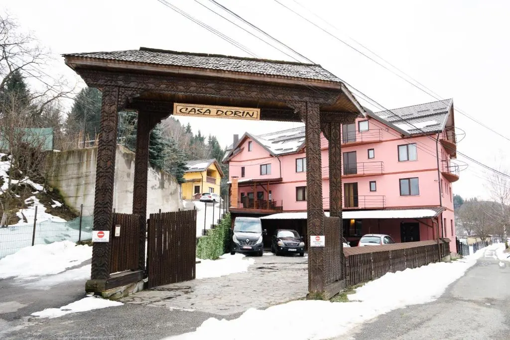 Pensiunea iarna: poarta de lemn, zăpadă pe margini și mașini parcate în curte.