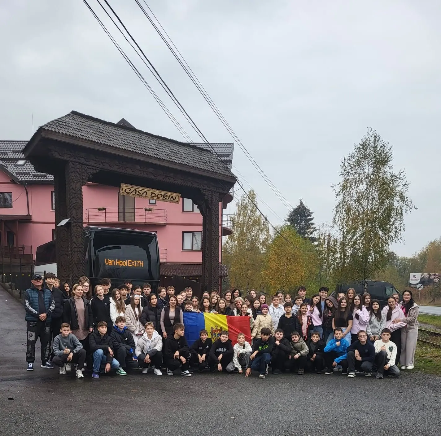 Grup de copii cu steagul României în fața pensiunii și autocarul în curte.