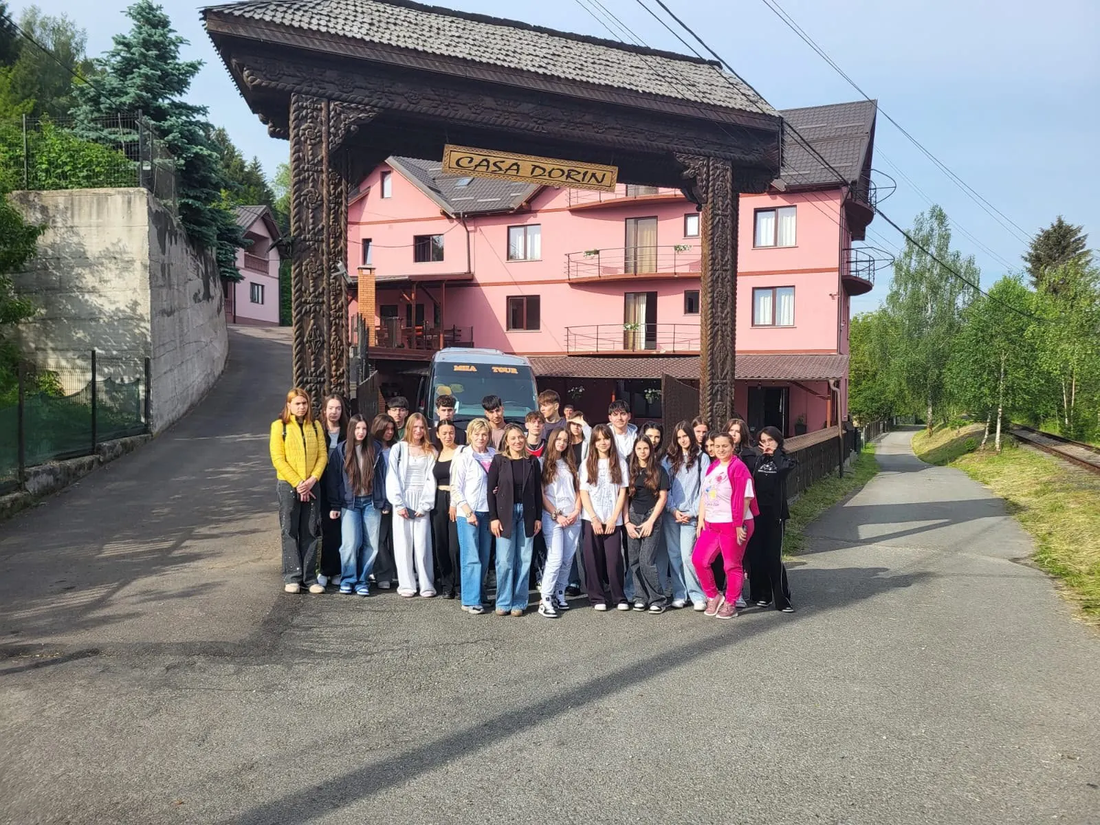 Grup de elevi în fața porții pensiunii, cu autobuzul în curte.