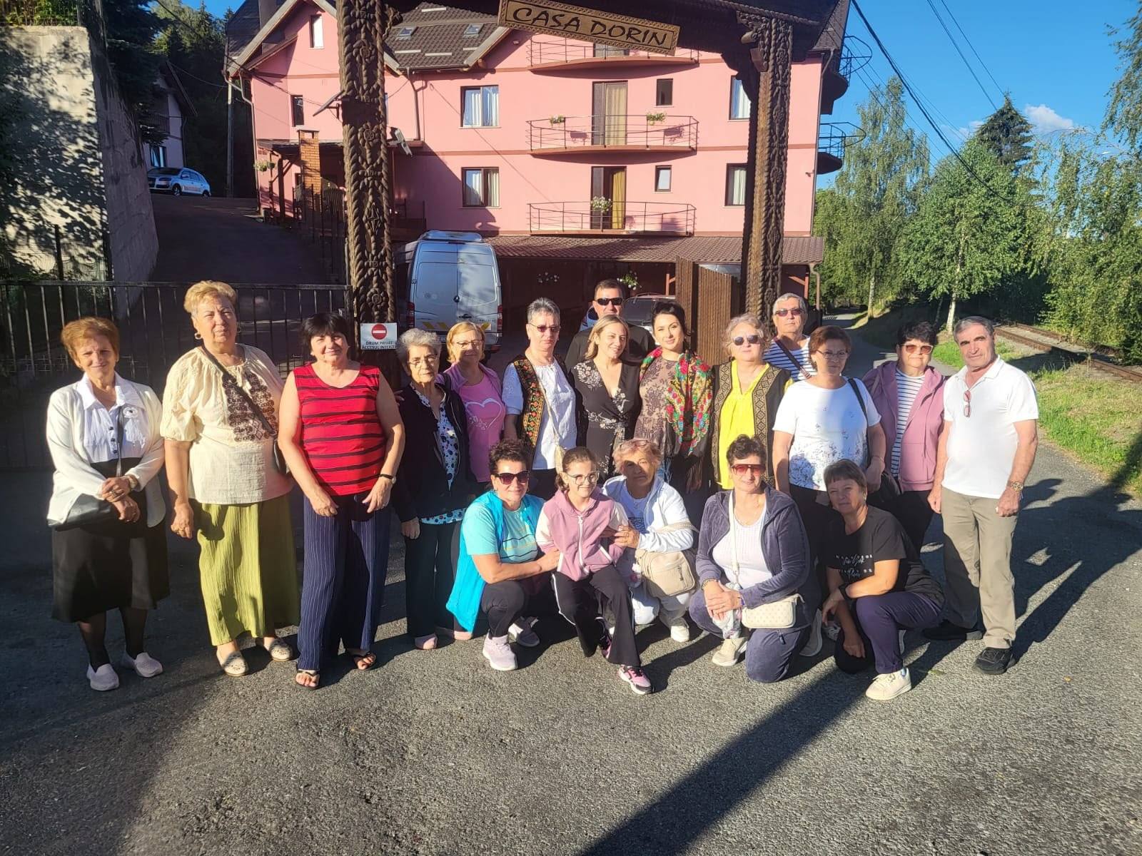 Grup de seniori fotografiați în fața porții pensiunii, într-o zi însorită.