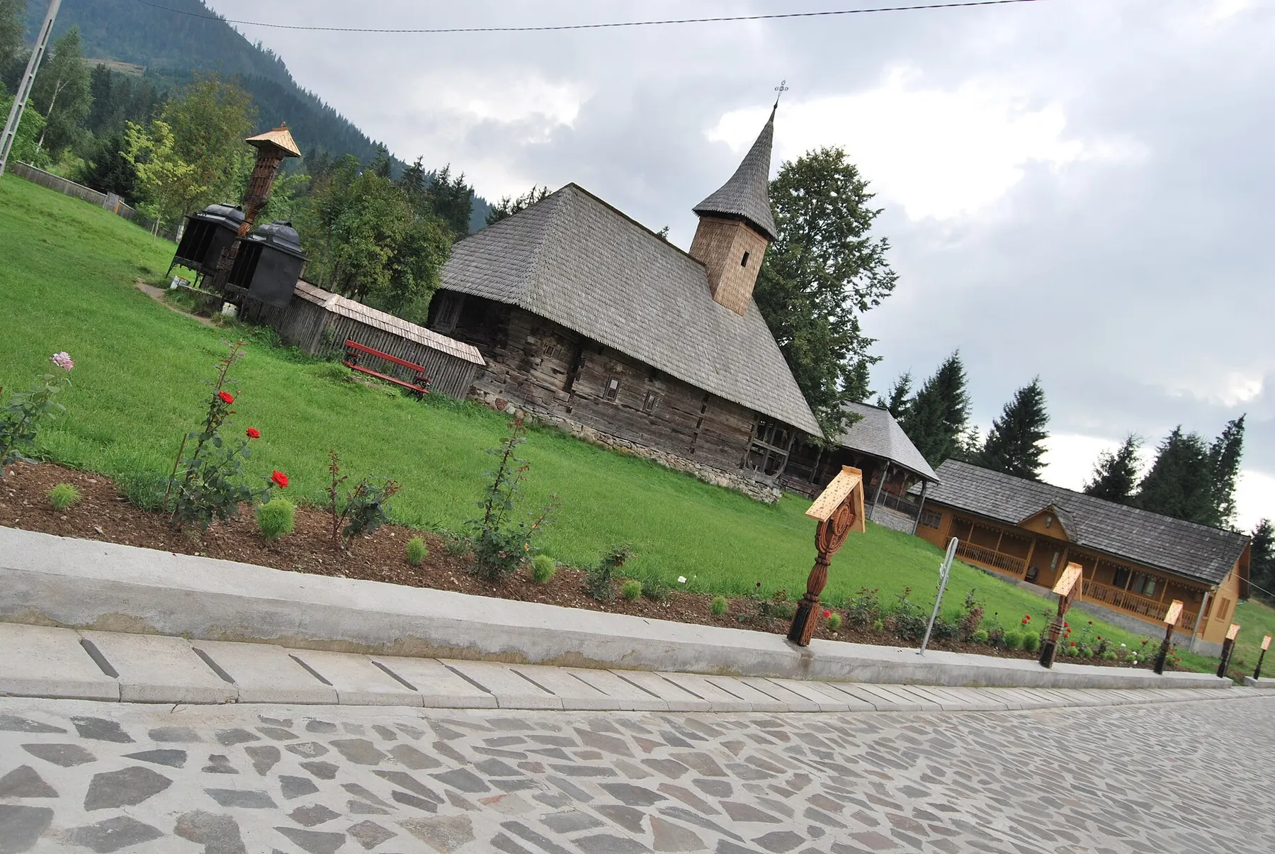 Biserica veche de lemn a Mănăstirii Moisei, cu acoperiș de șindrilă, pe un platou cu gazon și brazi.