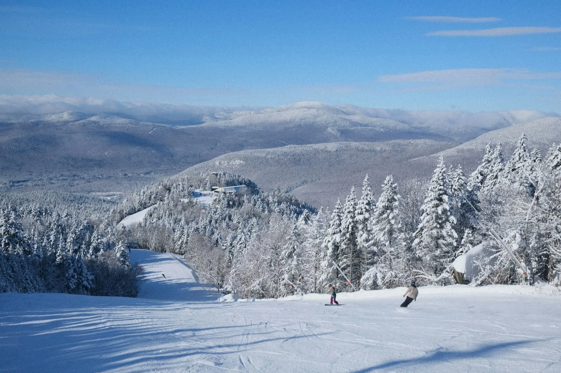 Pârtie de schi iarna, printre brazi încărcați de zăpadă, cu doi snowboarderi și munți în depărtare.