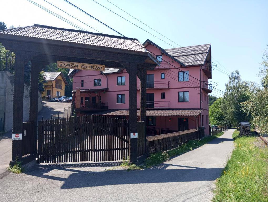 Poarta de lemn sculptat cu firma Casa Dorin și clădirea roz a pensiunii, vara, de pe drumul de acces.