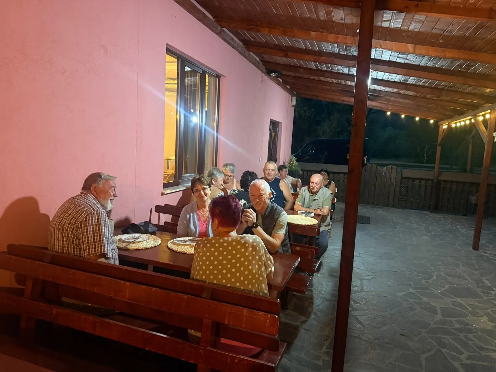 Oaspeți la cină pe terasa acoperită din curte, la mesele lungi de lemn.